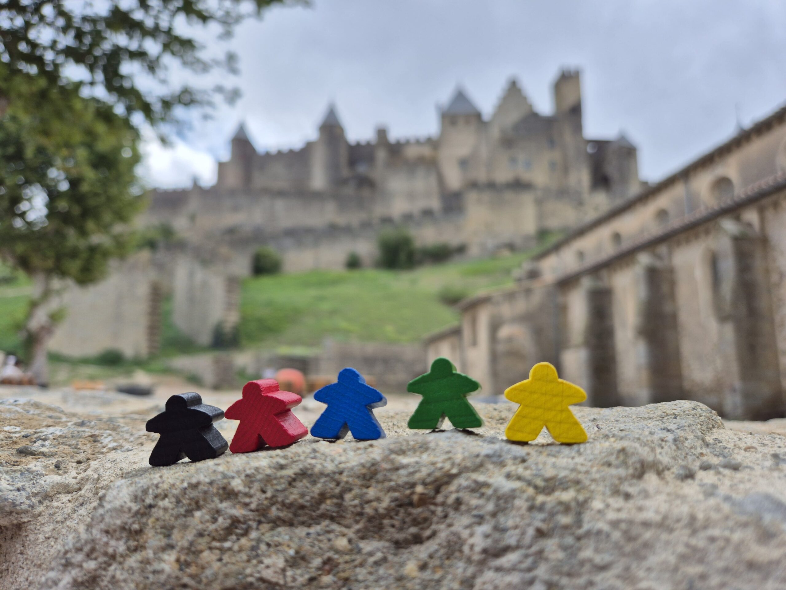 Carcassonne board game meeples on a stone wall with the medieval city of Carcassonne, France in the background.