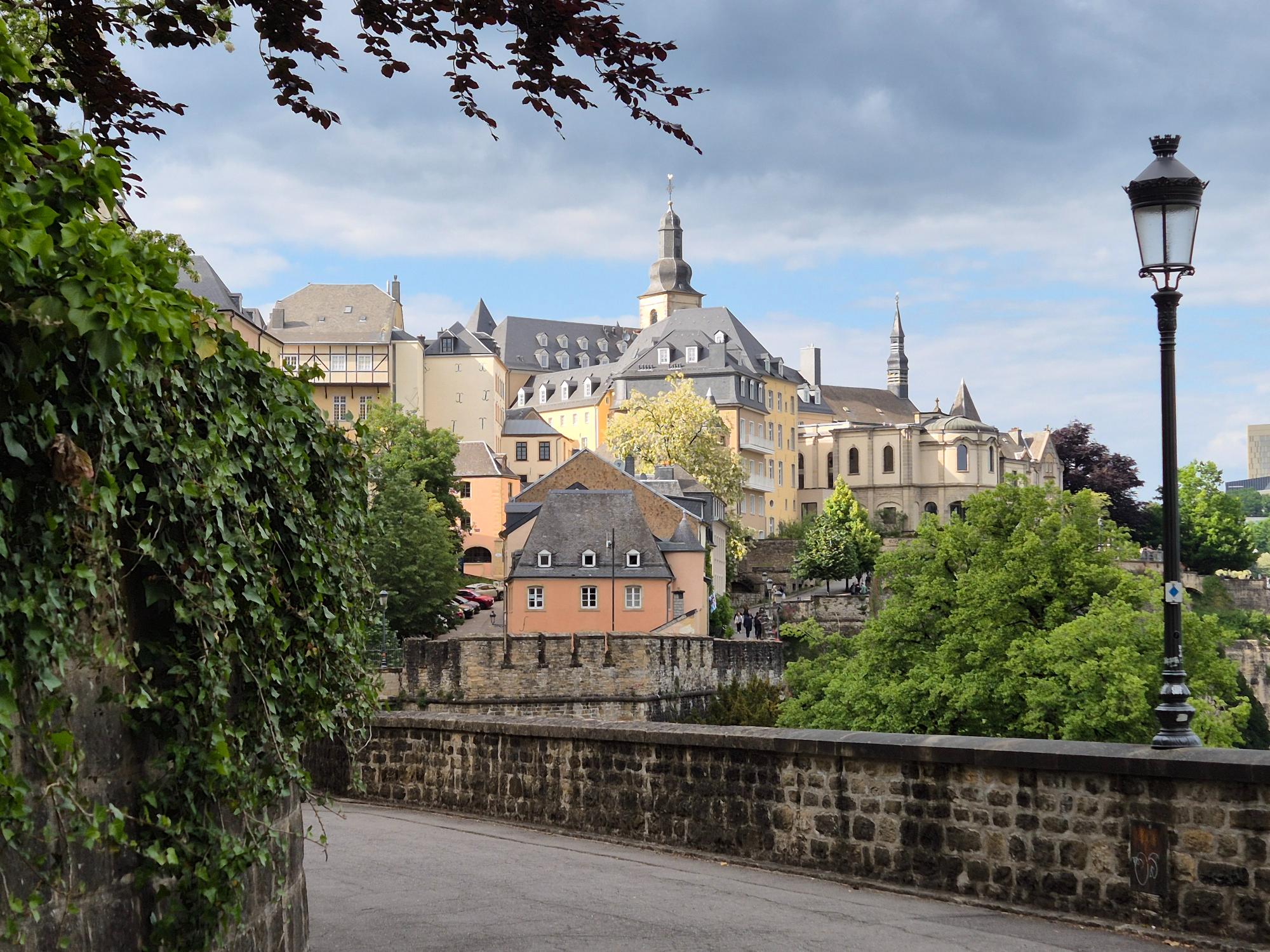 Historic stone ramparts and old town buildings in Luxembourg City with the spire of St. Michel Church under a cloudy sky.