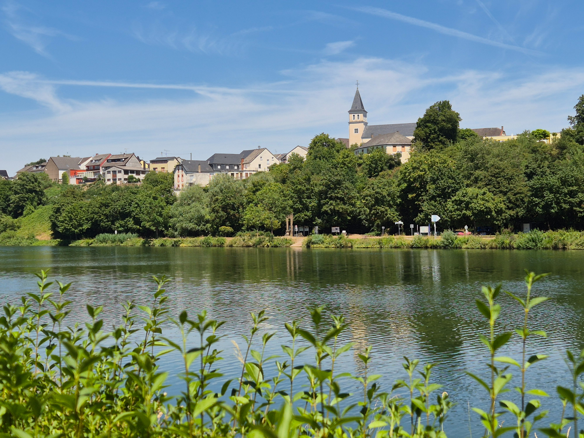 Lush green bushes framing a wide scenic view across the calm Sauer River toward hillside village houses and a historic church tower on the border of Luxembourg.
