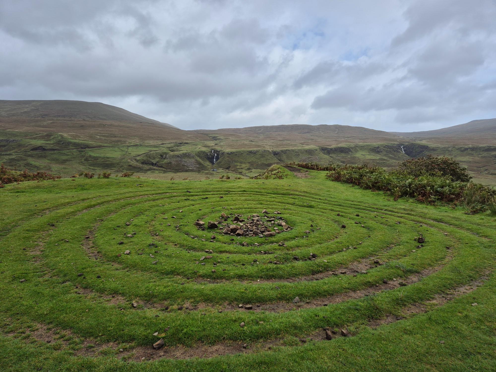 A circular green grass labyrinth with concentric paths winding inward to a stone center among rolling misty hills, symbolizing the journey into the subconscious mind.